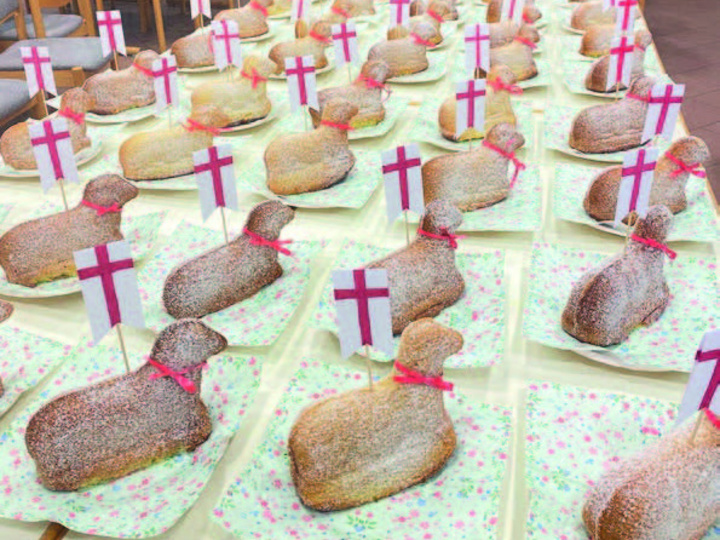 Das Bild zeigt einen Raum mit mehreren Tischen, auf denen längliche Brote in der Form eines Lammes liegen. Jedes Brot hat eine weiße Fahne mit einem roten Kreuz. Im Hintergrund sind Stühle und ein Kreuz an der Wand zu sehen.